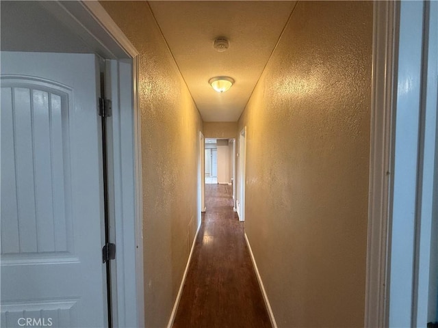 hallway featuring a textured wall, dark wood-style floors, and baseboards