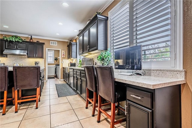 kitchen with light tile patterned floors, ornamental molding, decorative backsplash, under cabinet range hood, and plenty of natural light