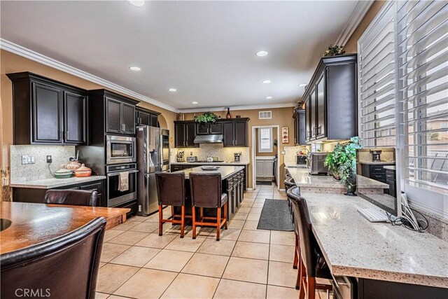 kitchen featuring appliances with stainless steel finishes, under cabinet range hood, crown molding, decorative backsplash, and a kitchen island