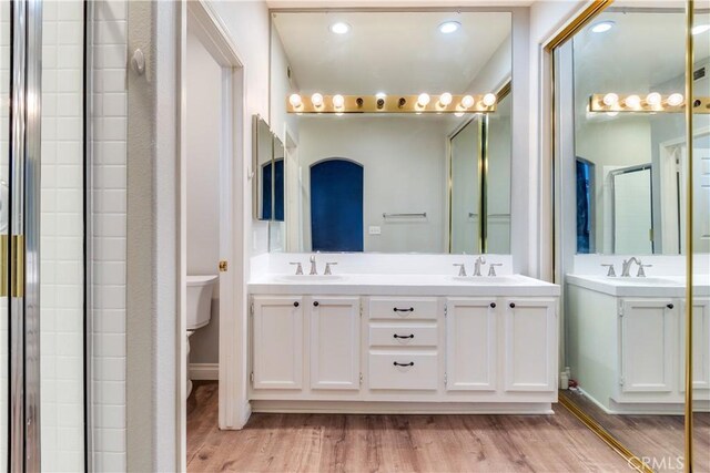 bathroom featuring wood finished floors, toilet, double vanity, and a shower with shower door