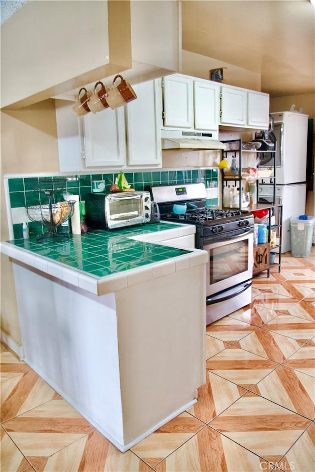 837 W 103rd St, Los Angeles, CA 90044 - kitchen featuring stainless steel gas stove, tile countertops, under cabinet range hood, freestanding refrigerator, and white cabinetry