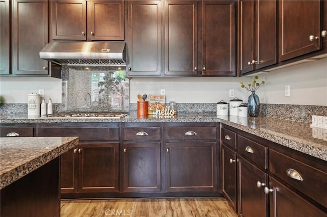 kitchen featuring under cabinet range hood, dark stone countertops, dark brown cabinets, and light wood finished floors