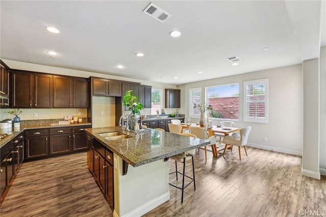 kitchen featuring a sink, dark wood-type flooring, dark stone countertops, an island with sink, and baseboards