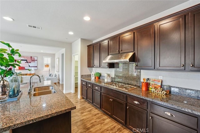 kitchen featuring under cabinet range hood, stainless steel gas cooktop, a sink, dark brown cabinetry, and dark stone counters