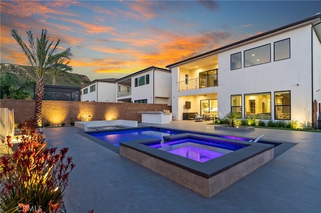 back of property at dusk featuring an in-ground hot tub, a balcony, a patio, and stucco siding