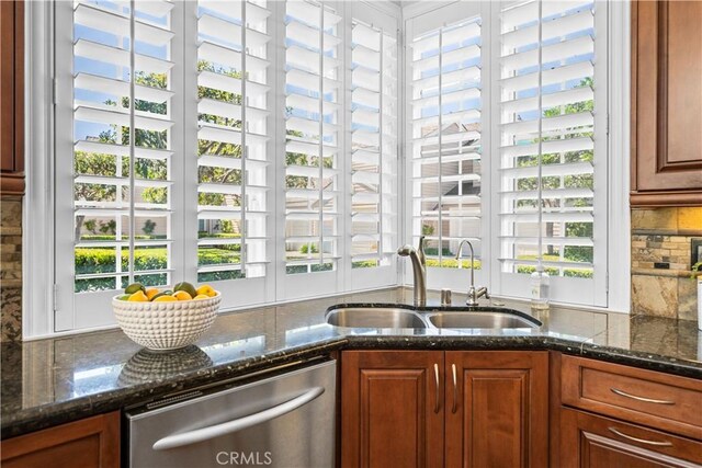 kitchen with stainless steel dishwasher, a sink, tasteful backsplash, dark stone counters, and brown cabinets