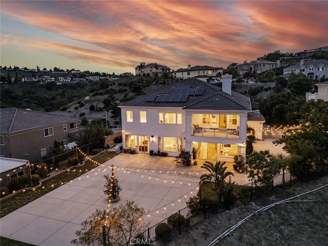 back of house at dusk with a balcony, solar panels, a fenced backyard, and a chimney