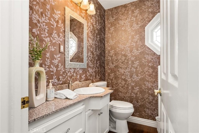 half bath with wood finished floors, toilet, baseboards, vanity, and wallpapered walls
