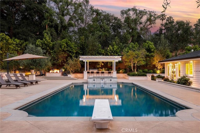 pool at dusk with a patio area, a diving board, an outdoor pool, view of wooded area, and a pergola