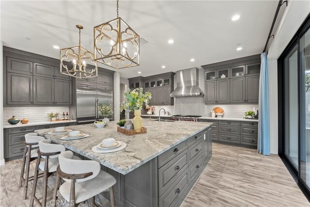 kitchen with wall chimney range hood, gray cabinets, stainless steel built in refrigerator, a chandelier, and decorative backsplash