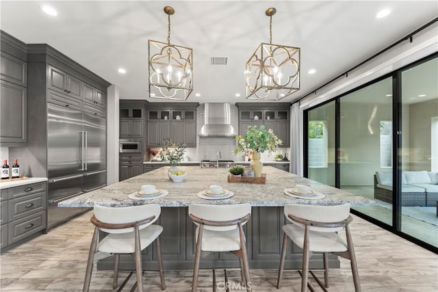 kitchen with stainless steel built in refrigerator, gray cabinetry, wall chimney exhaust hood, a chandelier, and backsplash