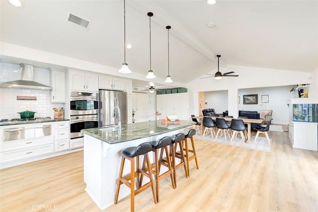 kitchen with wall chimney exhaust hood, a warming drawer, appliances with stainless steel finishes, a ceiling fan, and light wood-style flooring
