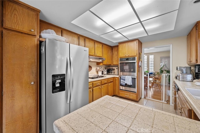 kitchen featuring appliances with stainless steel finishes, under cabinet range hood, light countertops, brown cabinetry, and light tile patterned flooring