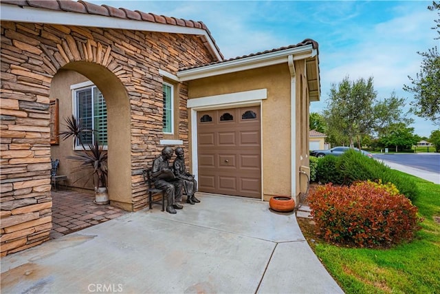 entrance to property with stone siding, an attached garage, stucco siding, and a tiled roof