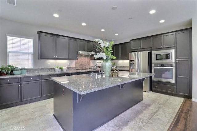kitchen featuring appliances with stainless steel finishes, under cabinet range hood, light stone counters, recessed lighting, and a kitchen breakfast bar