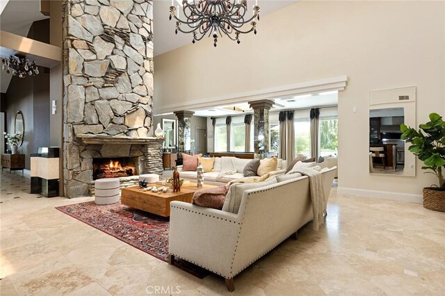 living room with a towering ceiling, a chandelier, ornate columns, a stone fireplace, and baseboards
