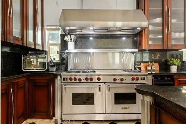 kitchen with double oven range, wall chimney range hood, tasteful backsplash, dark stone countertops, and glass insert cabinets
