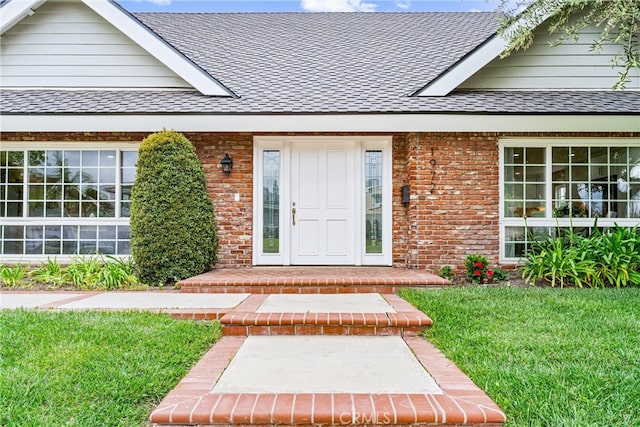 entrance to property with a shingled roof, a yard, and brick siding