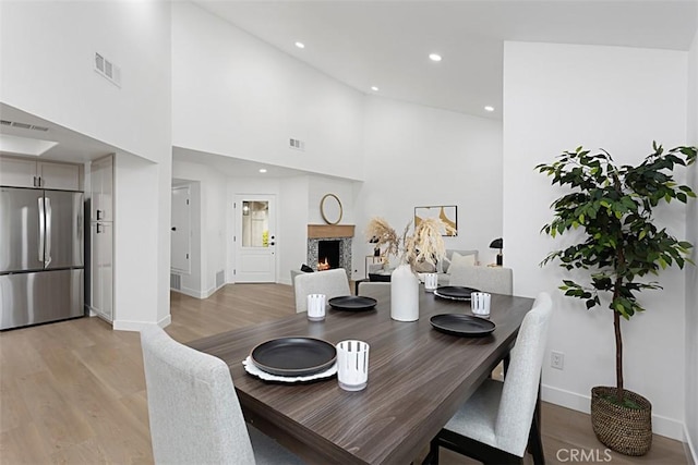 dining room featuring light wood finished floors, baseboards, a warm lit fireplace, recessed lighting, and a towering ceiling