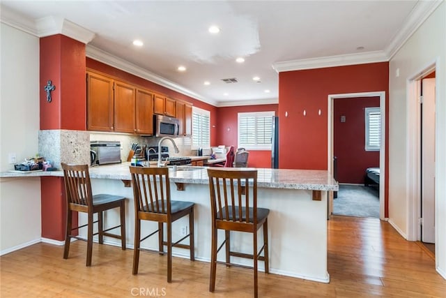 kitchen with a peninsula, stainless steel microwave, crown molding, light wood-type flooring, and brown cabinetry