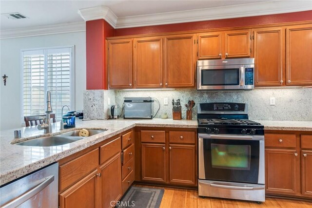 kitchen with stainless steel appliances, a sink, light stone countertops, brown cabinets, and ornamental molding