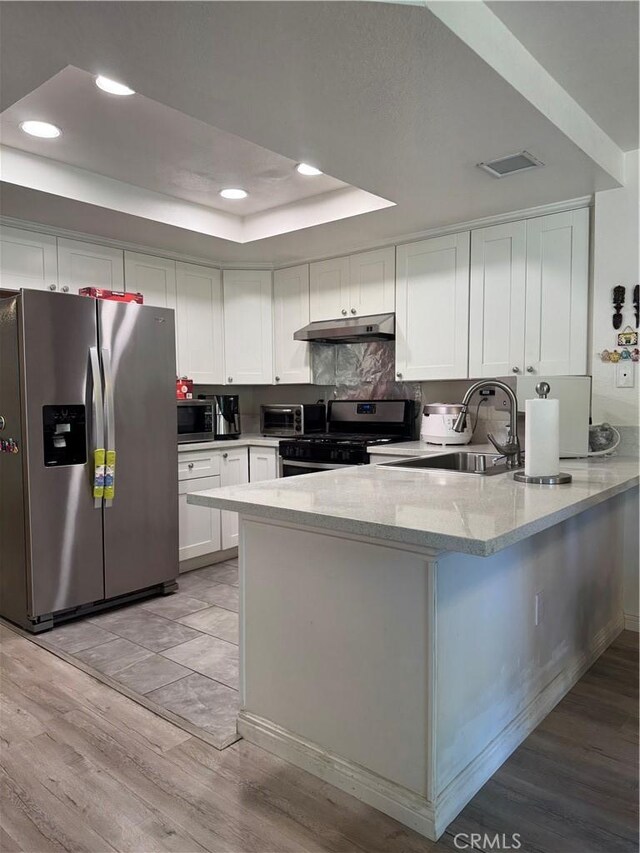 kitchen featuring stainless steel appliances, under cabinet range hood, a raised ceiling, a peninsula, and white cabinets