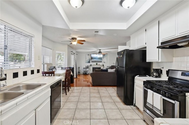 kitchen with black appliances, ceiling fan, under cabinet range hood, a tray ceiling, and a sink