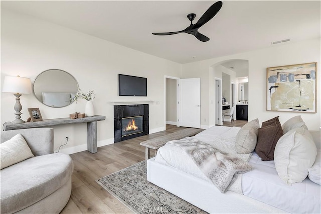 bedroom featuring arched walkways, a glass covered fireplace, wood finished floors, baseboards, and ensuite bath