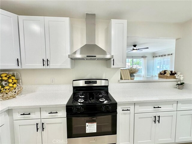kitchen with gas range, wall chimney exhaust hood, white cabinetry, ceiling fan, and light stone counters