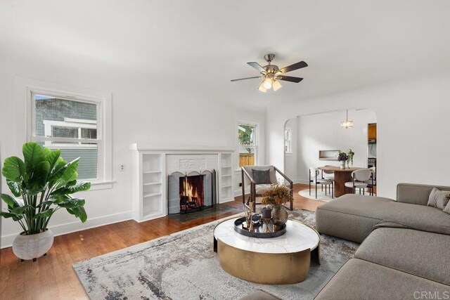 living area featuring baseboards, wood finished floors, a brick fireplace, a ceiling fan, and arched walkways