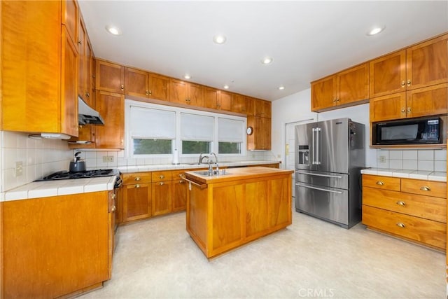 kitchen with black microwave, high end refrigerator, tasteful backsplash, under cabinet range hood, and a kitchen island with sink