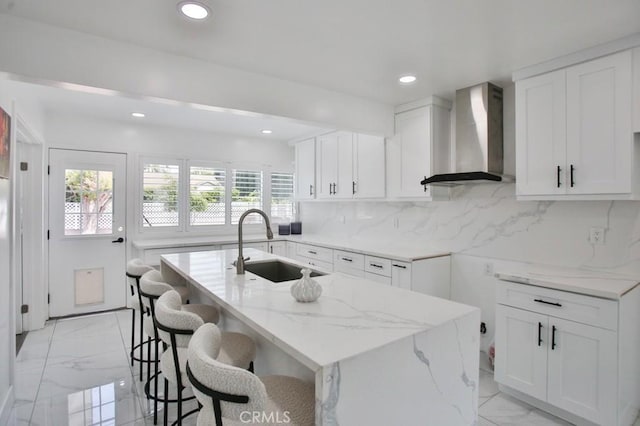 kitchen with wall chimney exhaust hood, a sink, light marble finish floors, light stone countertops, and recessed lighting