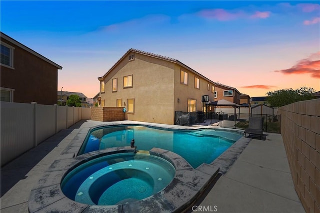 pool at dusk featuring a gazebo, a pool with connected hot tub, a fenced backyard, and a patio area
