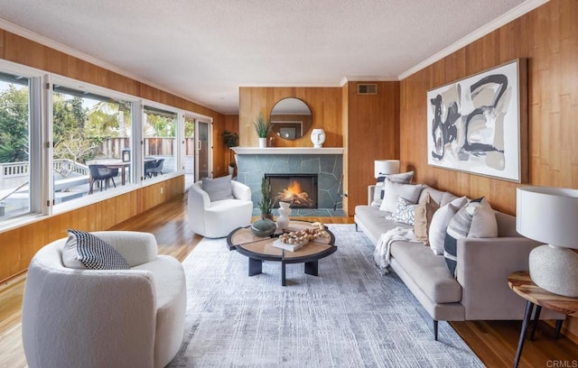 living room featuring wood walls, crown molding, wood finished floors, a stone fireplace, and a textured ceiling