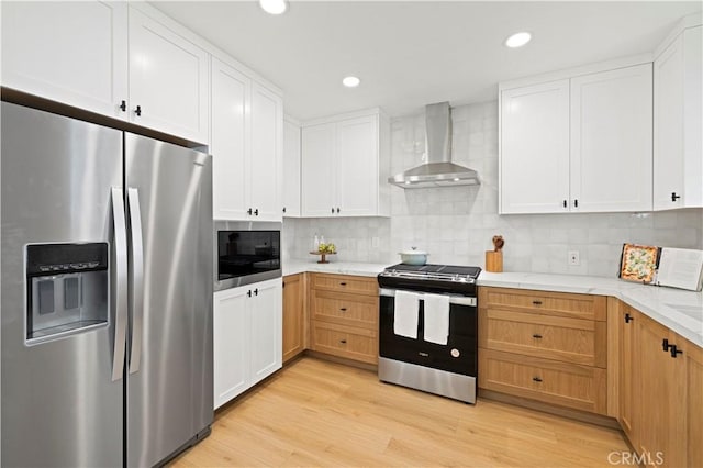 kitchen with appliances with stainless steel finishes, wall chimney range hood, light wood-style floors, backsplash, and white cabinetry