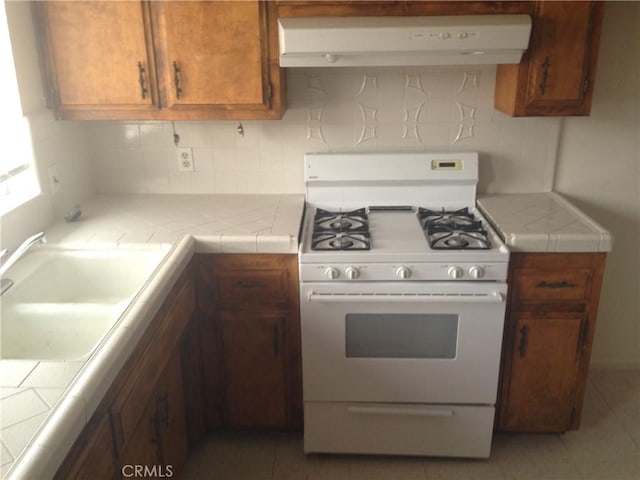 73601 Sun Valley Dr Unit 29, Palms, CA 92277 - kitchen featuring white range with gas stovetop, exhaust hood, tile counters, brown cabinetry, and tile patterned flooring