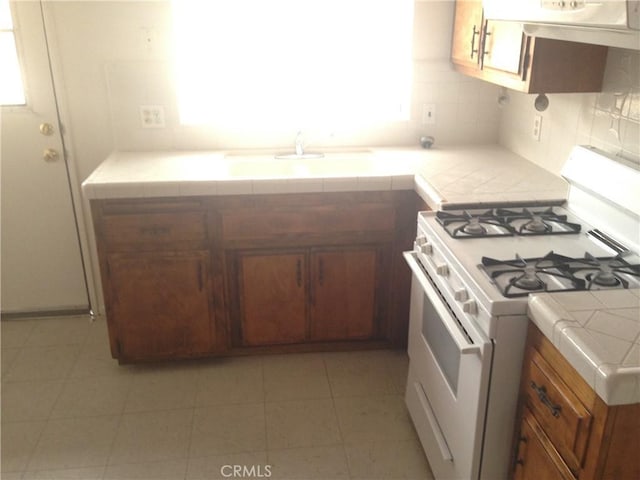 73601 Sun Valley Dr Unit 29, Palms, CA 92277 - kitchen with white range with gas stovetop, tile countertops, brown cabinets, range hood, and light tile patterned floors