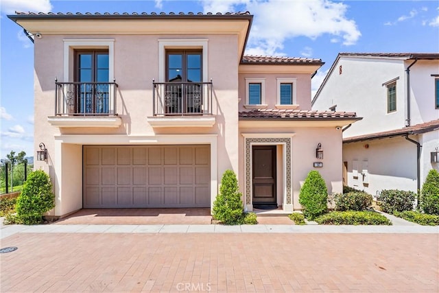 mediterranean / spanish house with a balcony, stucco siding, a tiled roof, a garage, and decorative driveway
