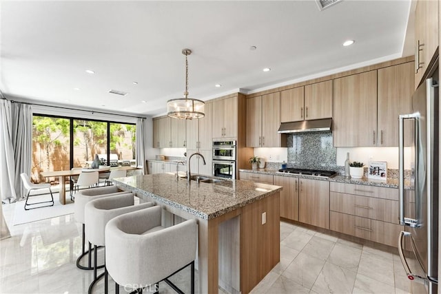kitchen featuring appliances with stainless steel finishes, under cabinet range hood, light brown cabinetry, a center island with sink, and a sink