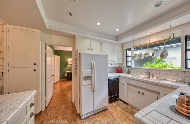 kitchen featuring tile countertops, white fridge with ice dispenser, black dishwasher, a tray ceiling, and recessed lighting