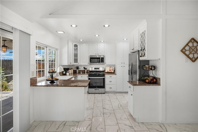 kitchen with appliances with stainless steel finishes, light marble finish flooring, a peninsula, dark stone counters, and a sink