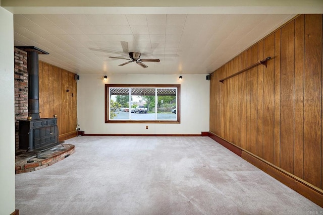 unfurnished living room featuring wooden walls, a wood stove, carpet, ceiling fan, and baseboards