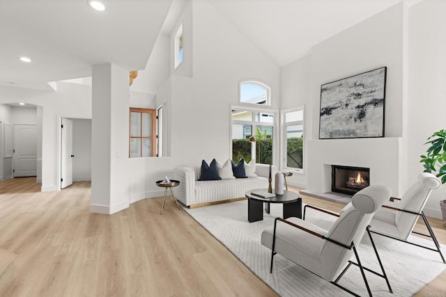 living room featuring a warm lit fireplace, light wood finished floors, high vaulted ceiling, baseboards, and recessed lighting