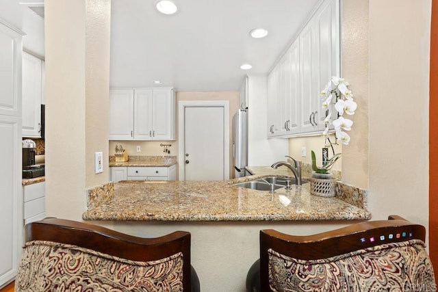 kitchen with a sink, freestanding refrigerator, a kitchen breakfast bar, light stone counters, and recessed lighting