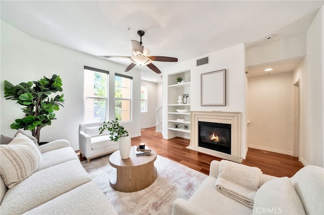 living room with built in shelves, a ceiling fan, wood finished floors, and baseboards