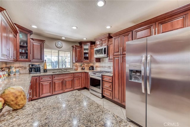 kitchen with appliances with stainless steel finishes, a sink, backsplash, light stone counters, and glass insert cabinets