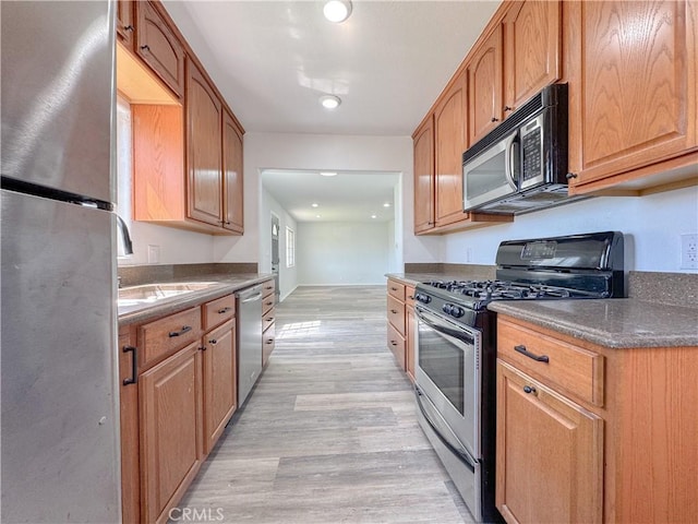 kitchen with appliances with stainless steel finishes, light wood-type flooring, dark countertops, a sink, and recessed lighting