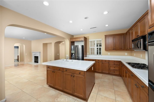 kitchen featuring appliances with stainless steel finishes, light tile patterned floors, a kitchen island with sink, light countertops, and brown cabinetry