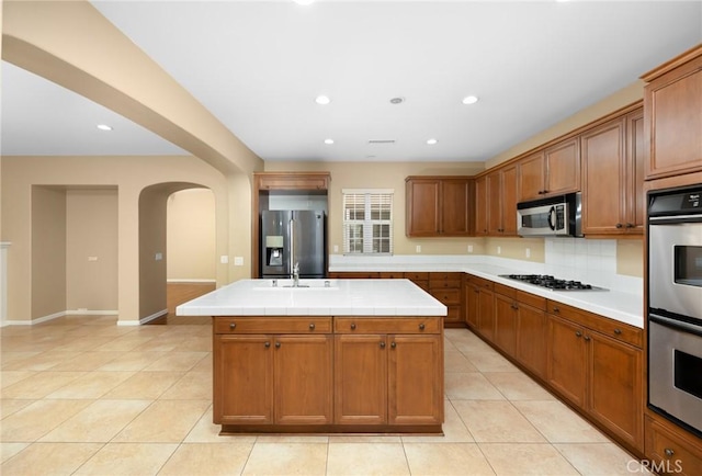 kitchen with stainless steel appliances, arched walkways, a kitchen island, light tile patterned floors, and brown cabinetry