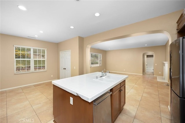 kitchen with arched walkways, a sink, freestanding refrigerator, light tile patterned floors, and an island with sink